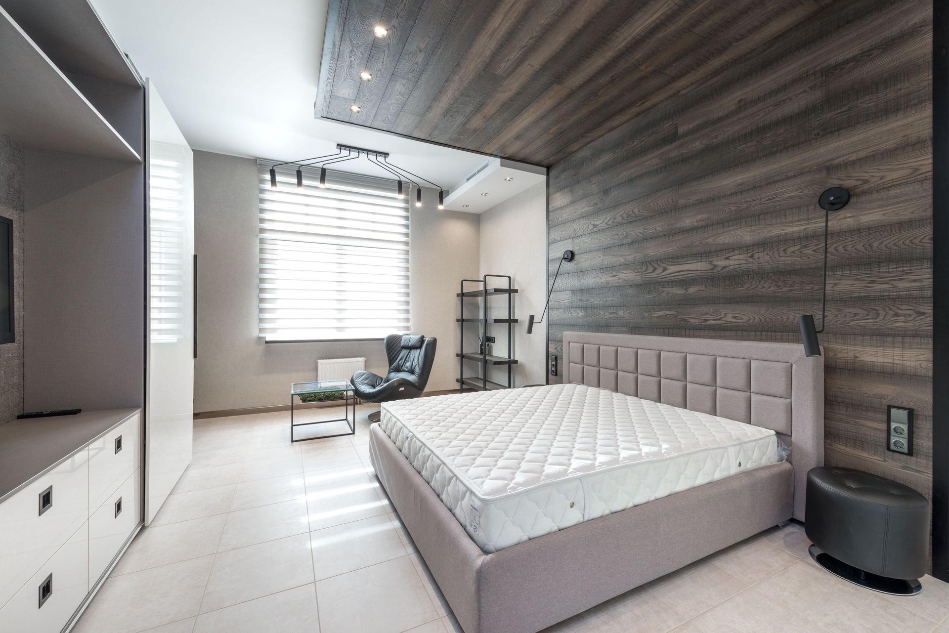 Bed with white mattress placed at wall with decorative element against cupboards in light stylish bedroom with window and armchair